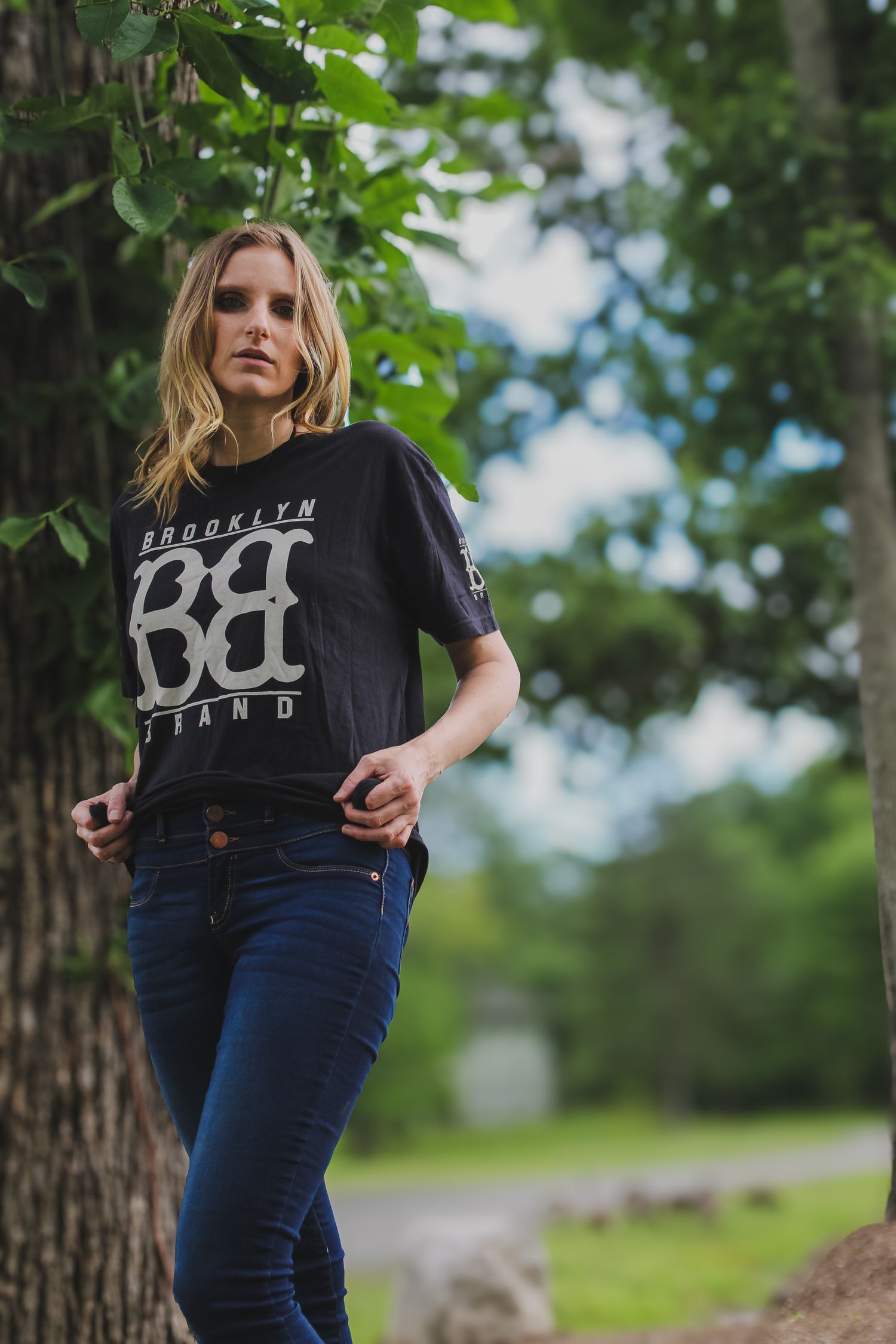 Person in a black graphic t-shirt and blue jeans poses casually against a tree trunk in an outdoor nature setting.