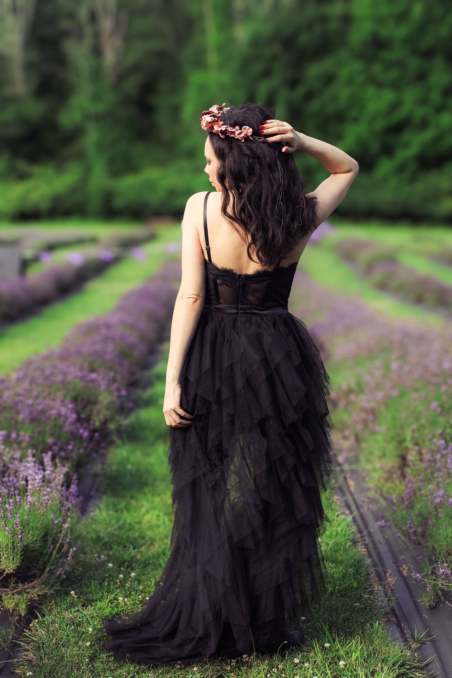 A figure in a flowing black dress poses gracefully among rows of blooming purple lavender flowers in a garden setting.