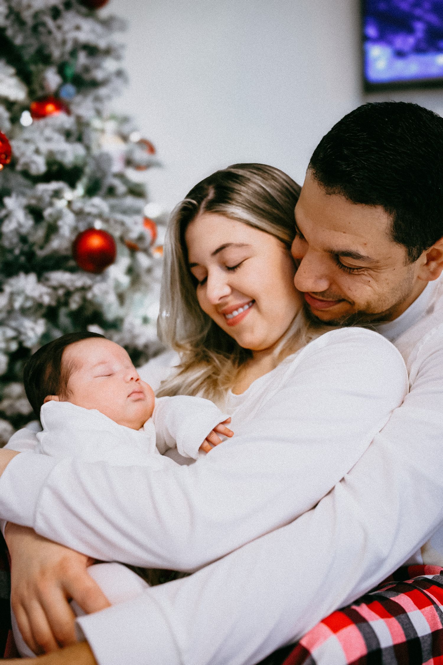 A couple in white snuggles together on a couch near a decorated Christmas tree with twinkling lights.