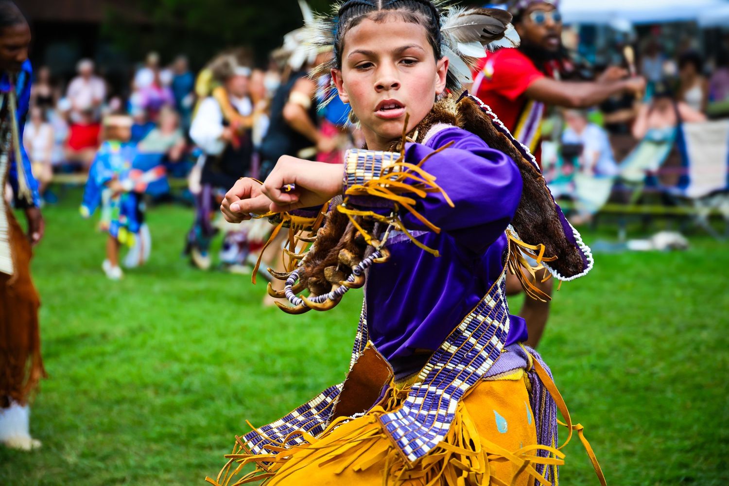 Native American dancers in traditional regalia perform at an outdoor powwow celebration on grassy field.