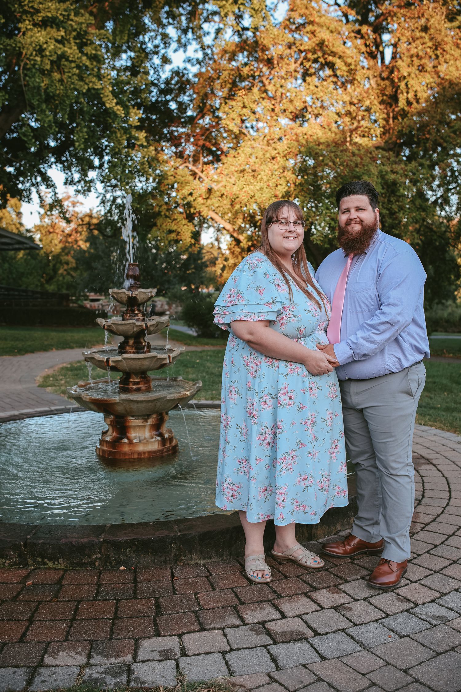 A couple poses together on a brick patio near a fountain surrounded by autumn trees with golden leaves.