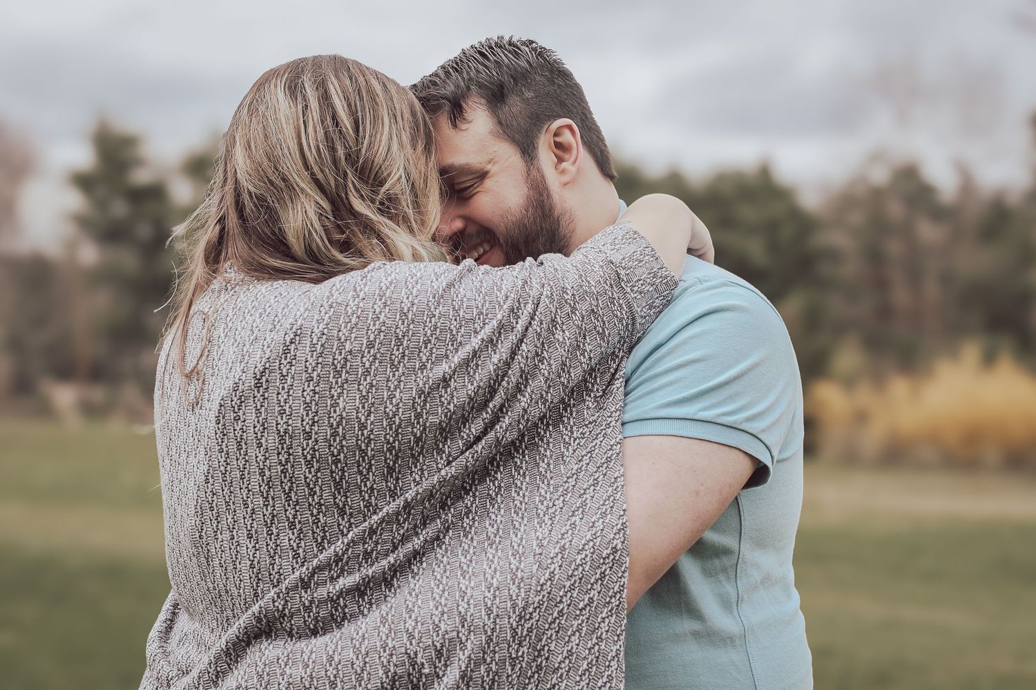 A couple embraces tenderly in a field during a cloudy autumn day while wearing cozy sweaters.