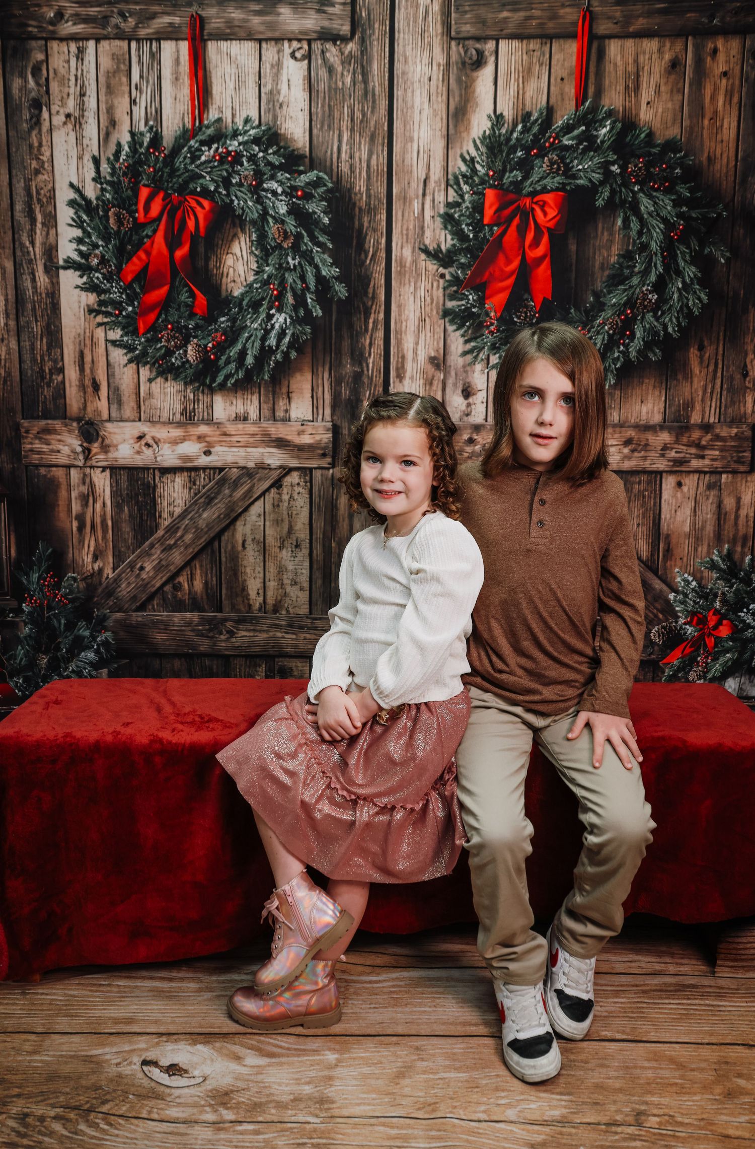 Children sitting on a red bench against rustic wooden wall decorated with Christmas wreaths and red ribbons.