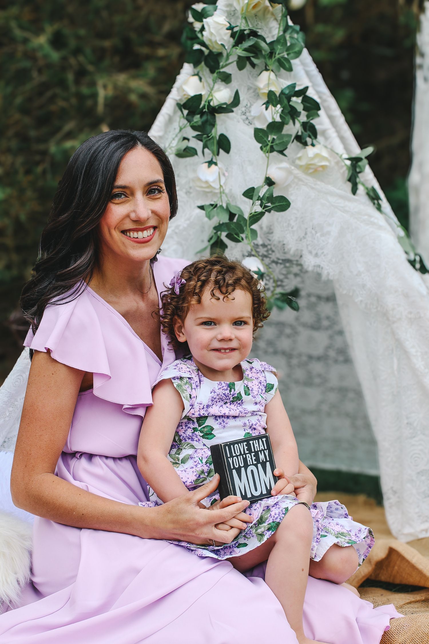 Mother and child in coordinating pink and floral outfits sit together in front of a decorated white teepee backdrop.