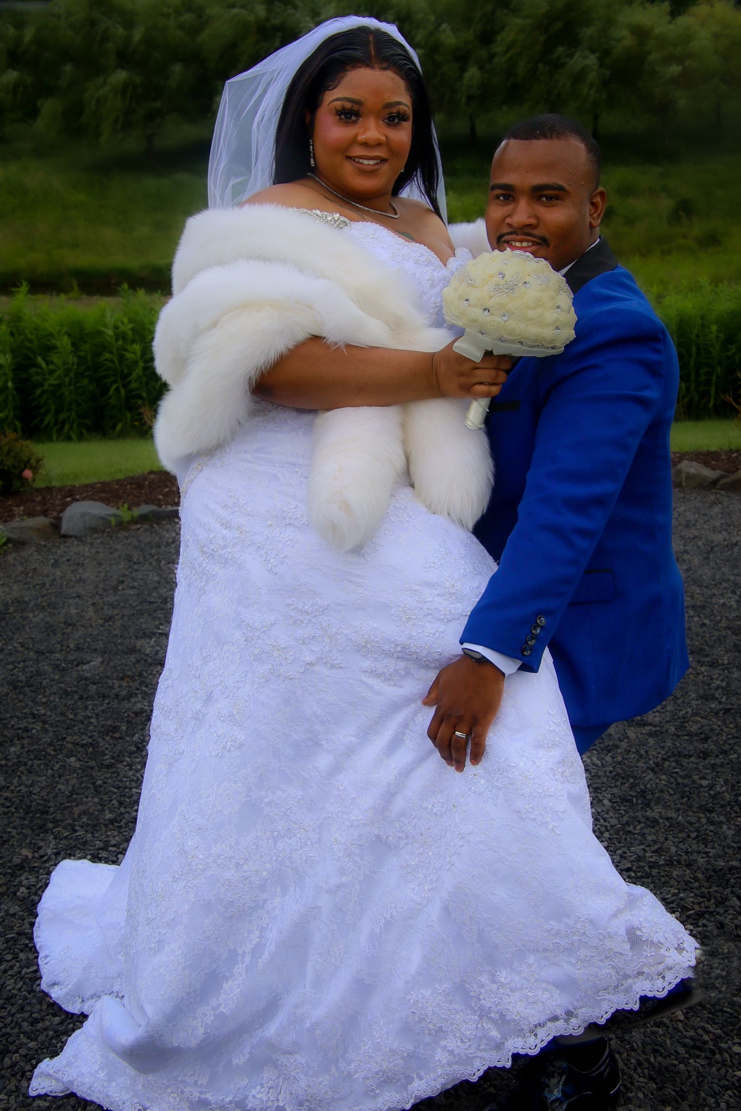 A couple poses together on their wedding day, with the bride in a white lace gown and fur wrap holding a bouquet.