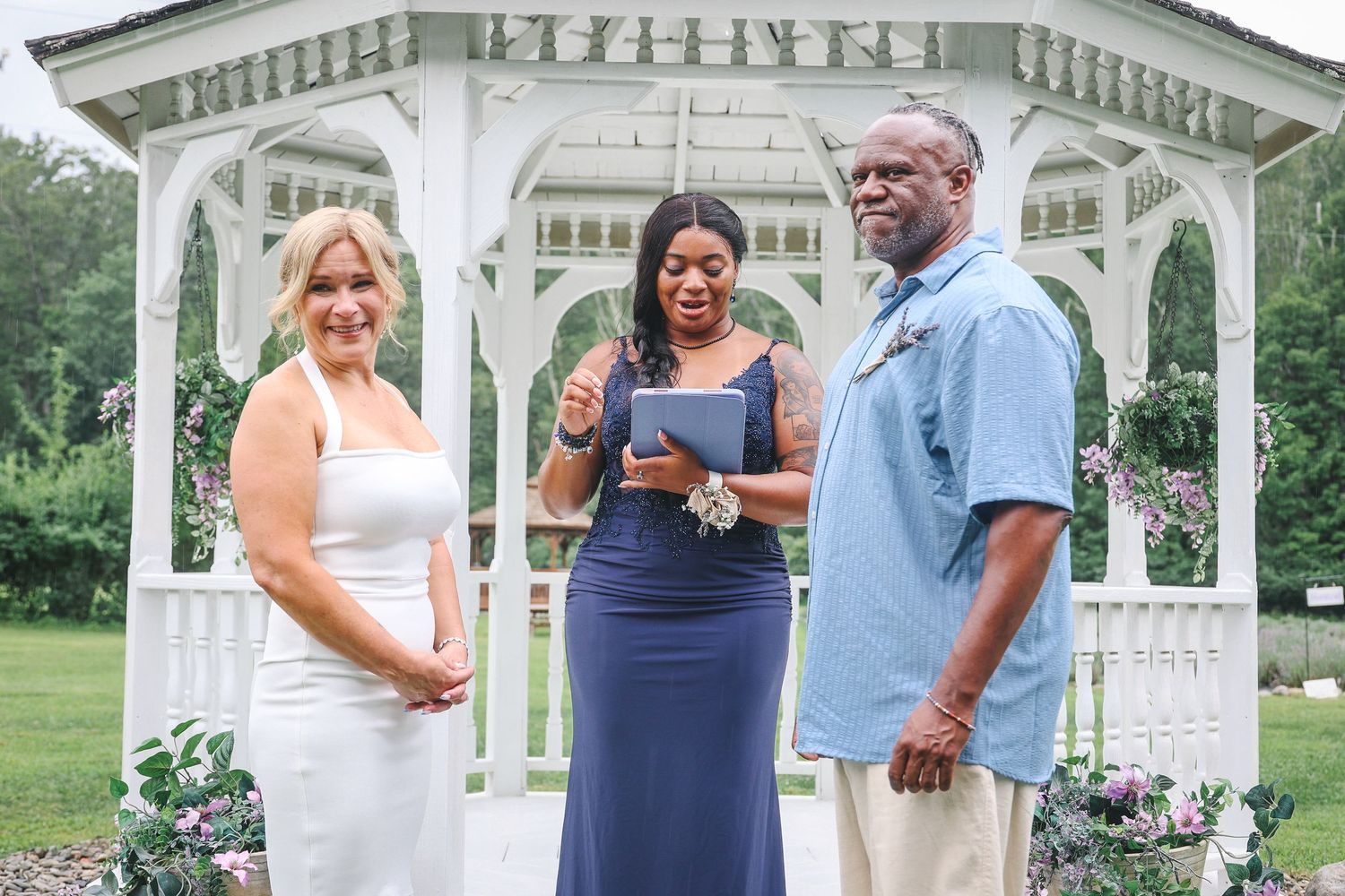 Wedding party poses in front of a white gazebo decorated with purple flowers on a sunny summer day.