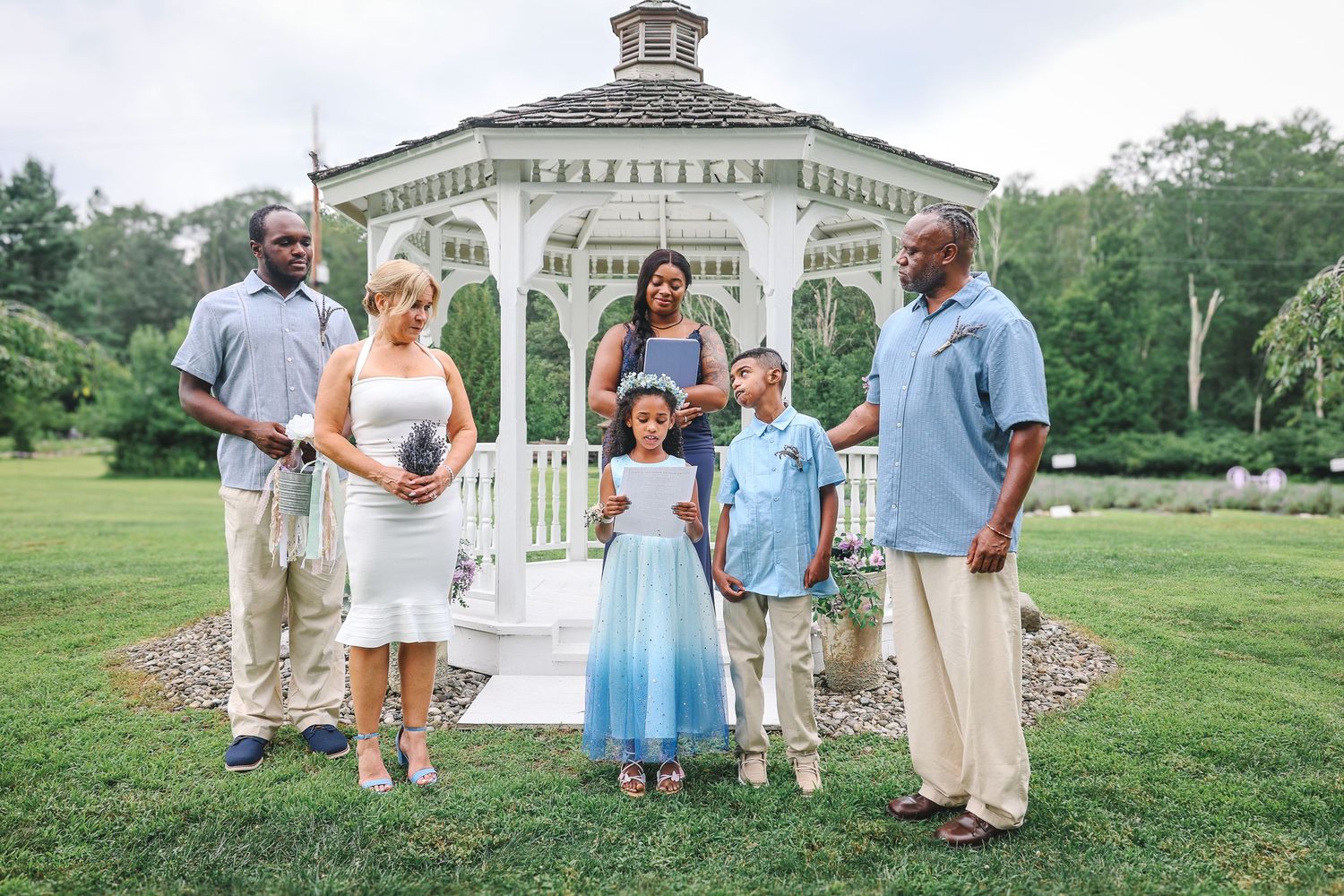 Wedding party members gather outdoors near a white gazebo and garden for formal photos in light blue and khaki attire.