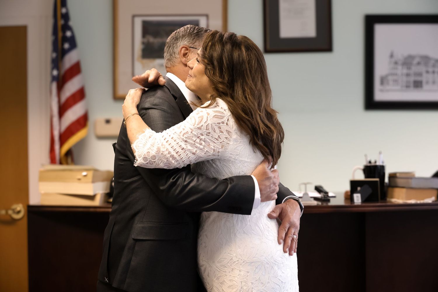 A couple in formal attire dances together in a room with American flags and framed photos on the wall.