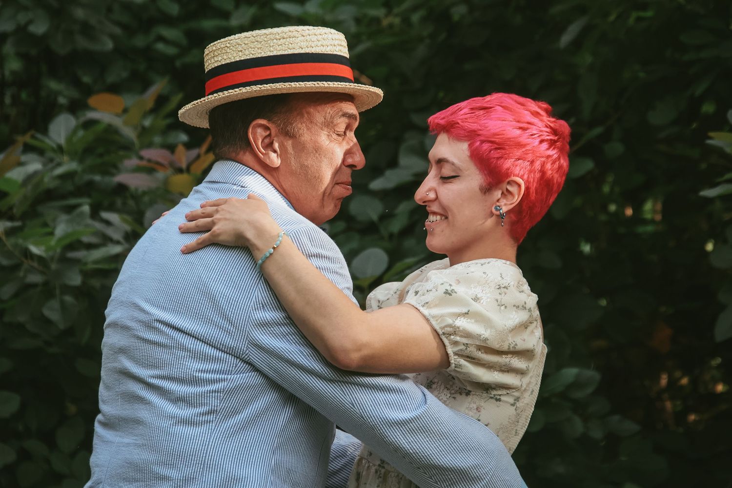 A couple embraces outdoors, with one wearing a straw boater hat and the other sporting vibrant pink hair.