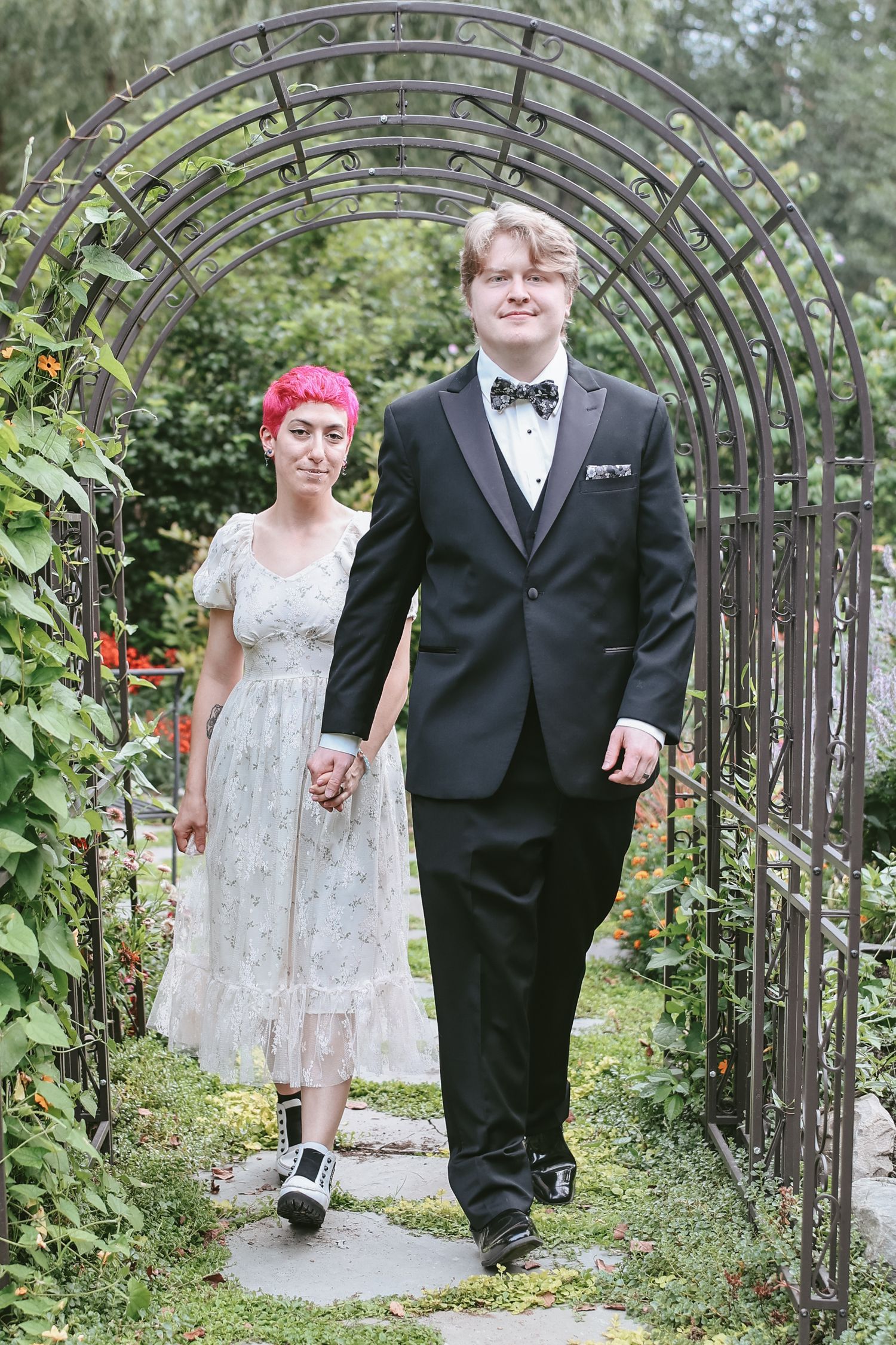 A couple walks through a garden archway, with the bride wearing a lace tea-length dress and the groom in a classic tuxedo.