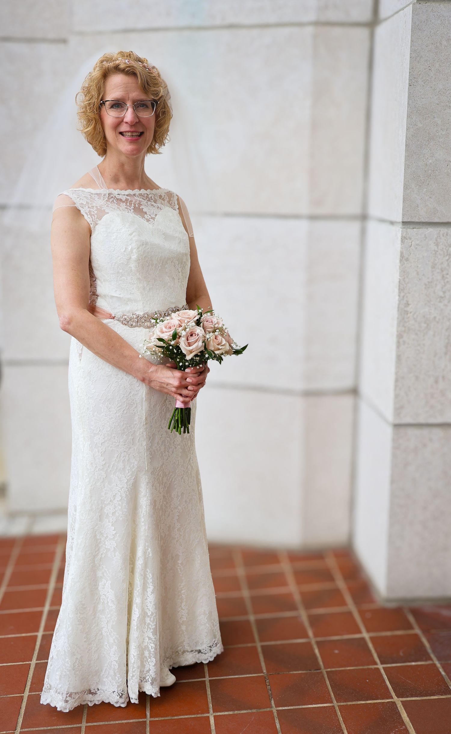 A bride in a white lace tea-length wedding dress poses with a pink bouquet on a terracotta tile floor.