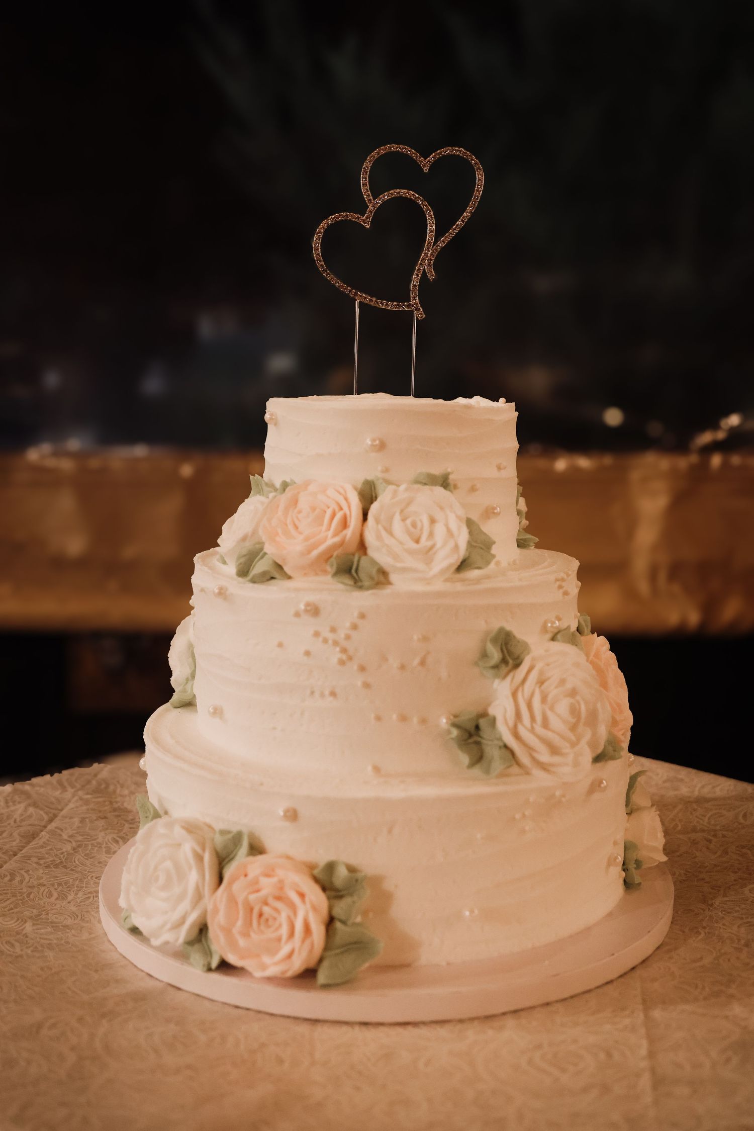 A three-tiered white wedding cake decorated with delicate pink roses and green leaves against a dark background.