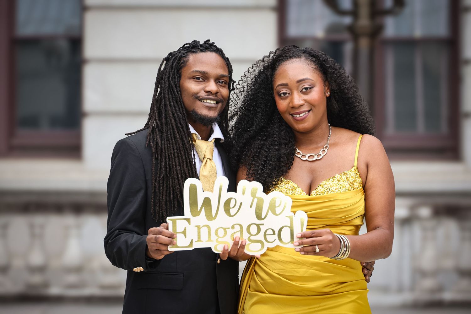 A couple in formal attire poses together, with the person in a bright yellow dress holding an engagement sign.