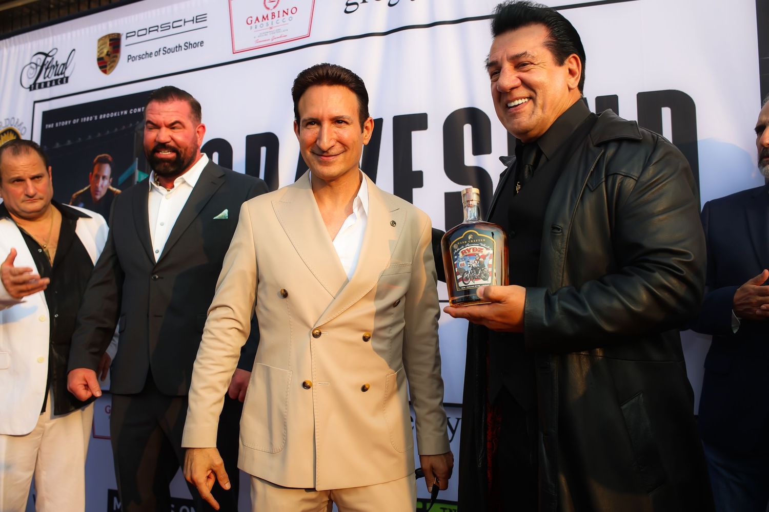 Three people stand together at a promotional event, with one wearing a beige suit holding a bottle of spirits.