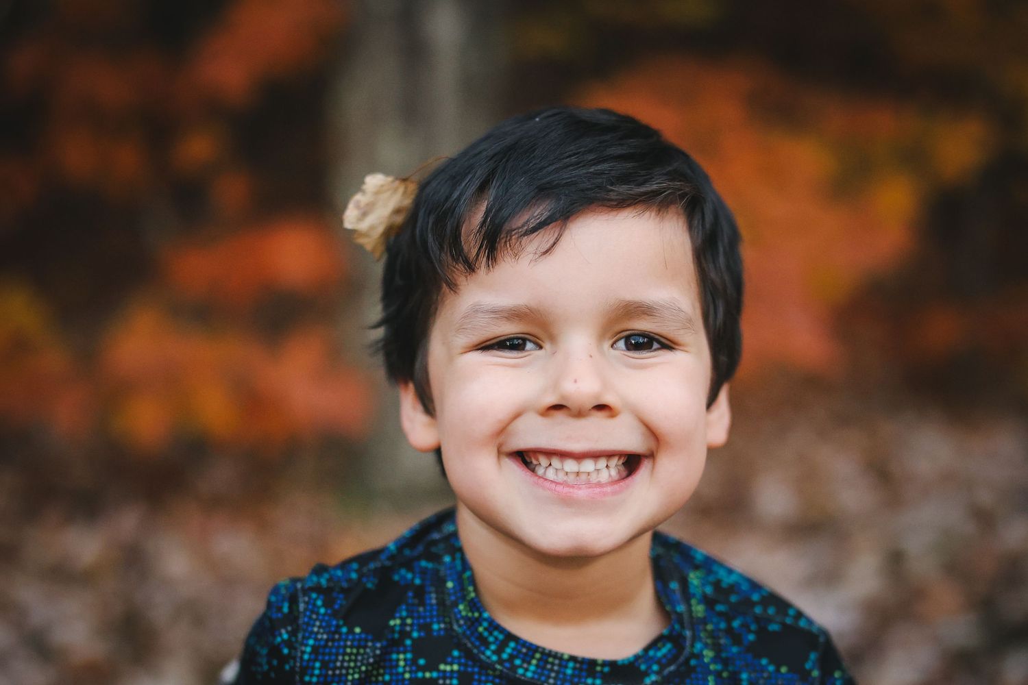 A young child smiles brightly at the camera while wearing a blue sweater against a blurred autumn background.