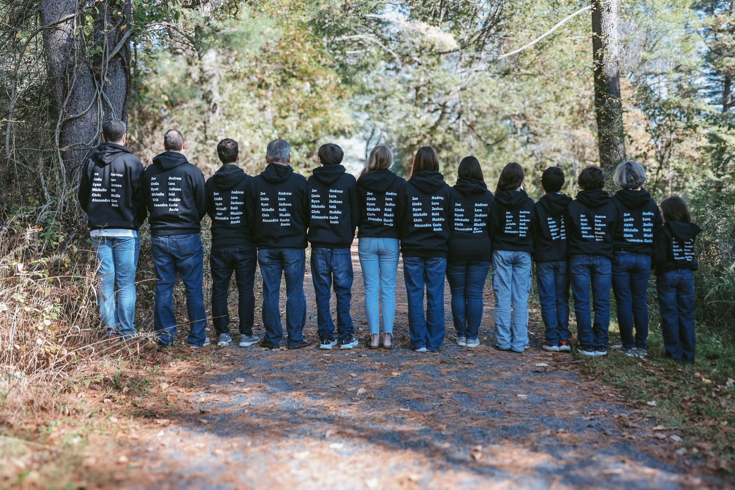 A group of people wearing matching black hoodies stand in a line with their backs turned on a wooded trail.