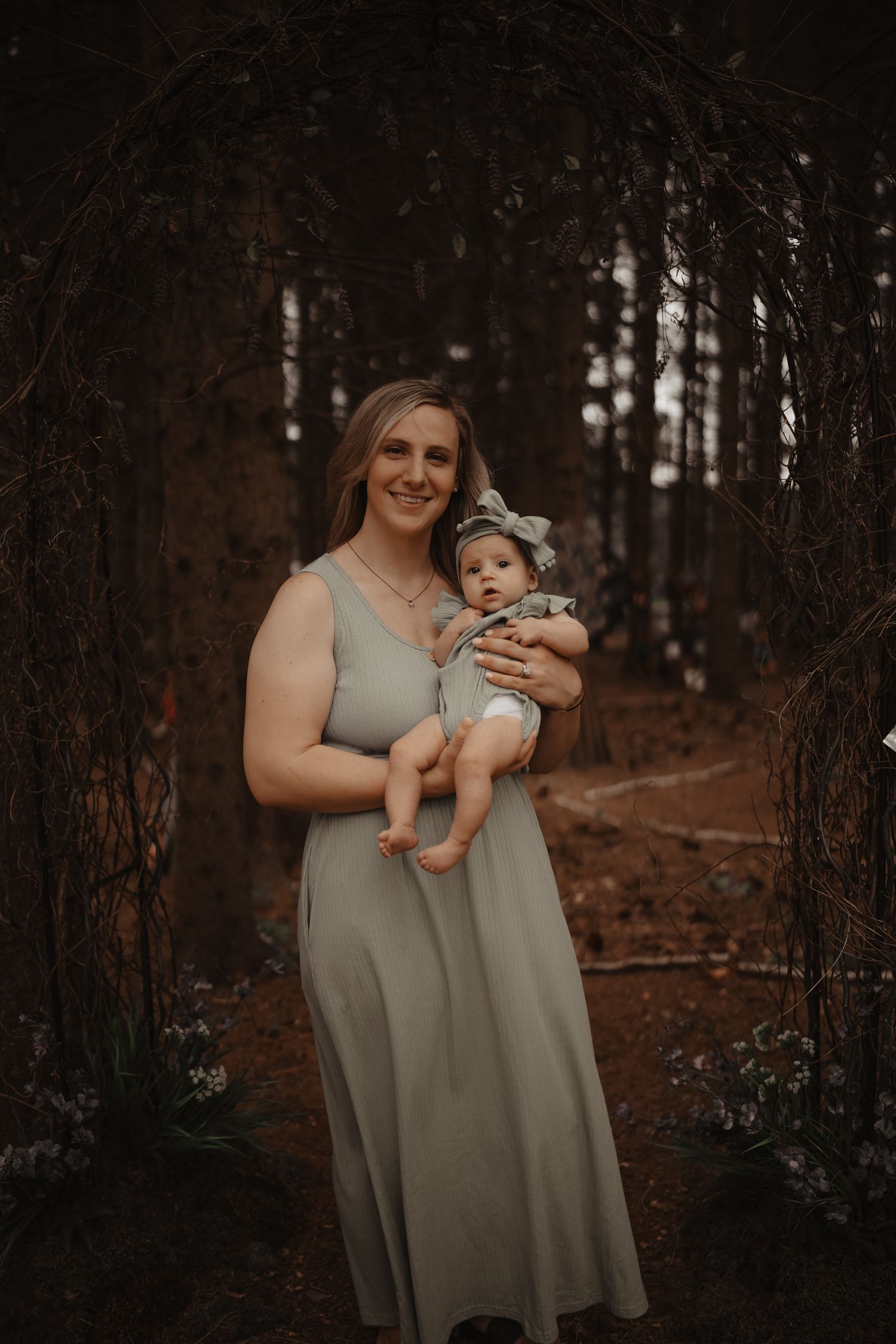A parent in a sage green dress holds a baby in a wooded setting during golden hour.