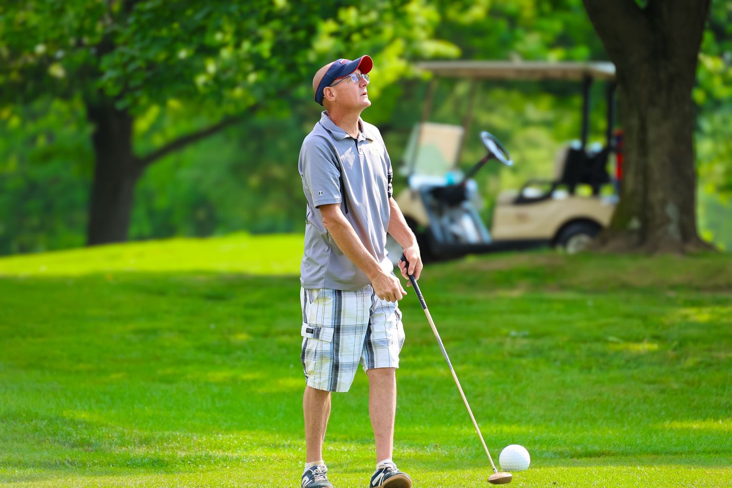 A golfer in plaid shorts and polo shirt stands ready to putt on a lush green course with a golf cart in background.