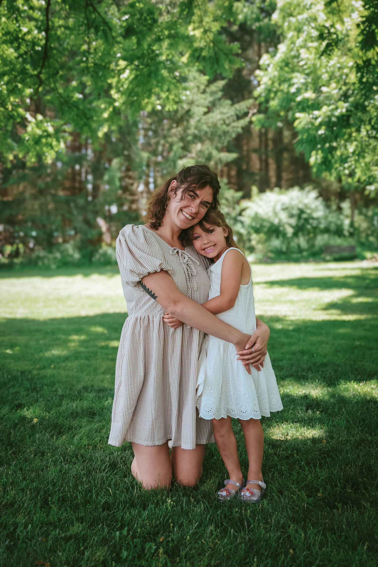 A woman kneeling on green grass hugging a young girl in a white dress outdoors in a lush garden.