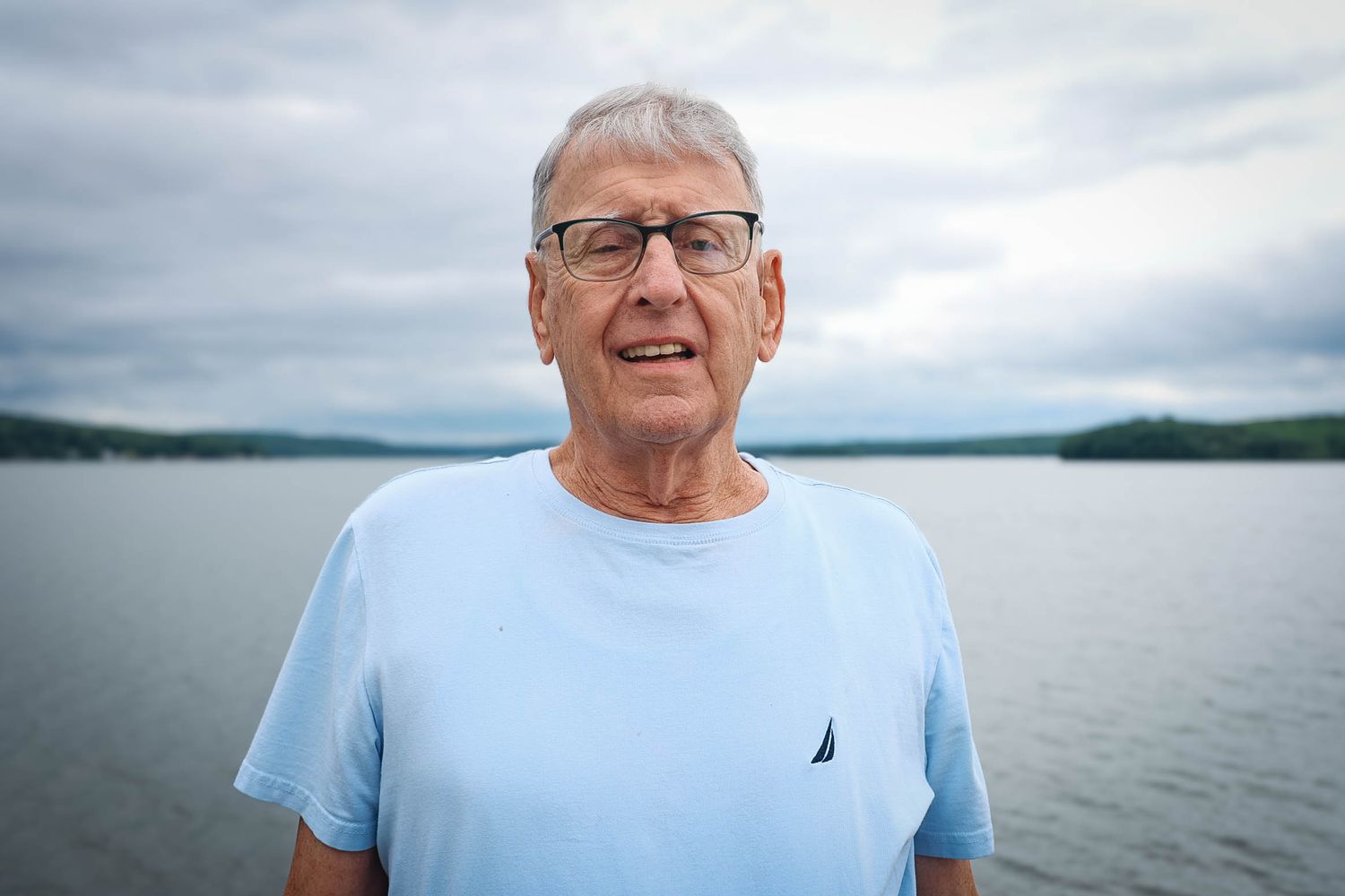 Elderly man in light blue Nautica t-shirt standing outdoors near a lake with cloudy skies in background.