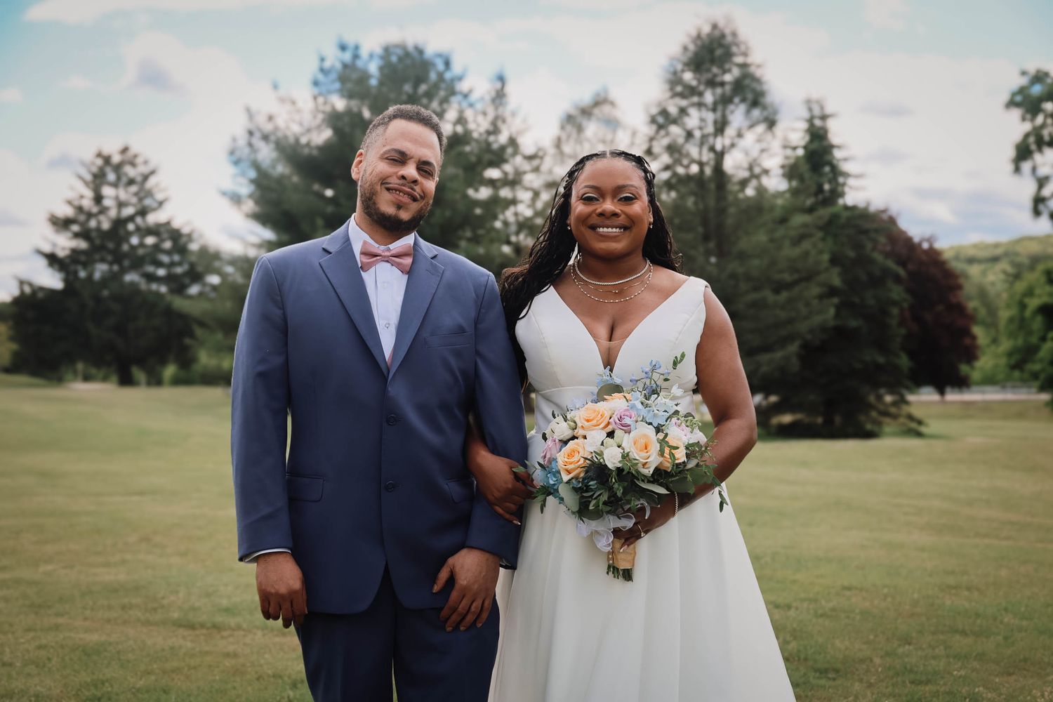 Bride and groom smiling outdoors on green lawn, bride holding pastel bouquet, groom in blue suit with pink bow tie.