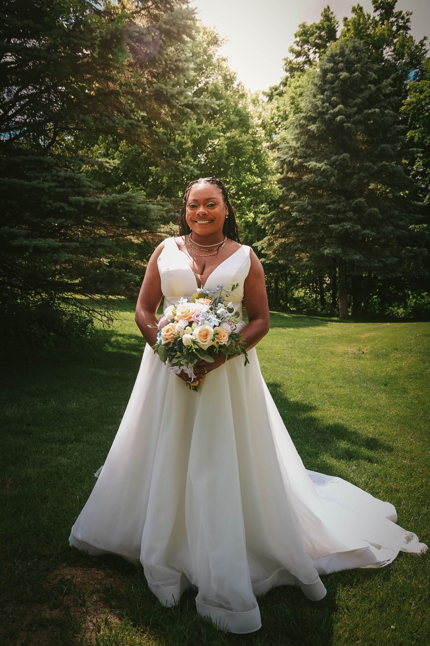 A smiling bride in a white ball gown holds a pastel bouquet while standing on green grass surrounded by lush trees.