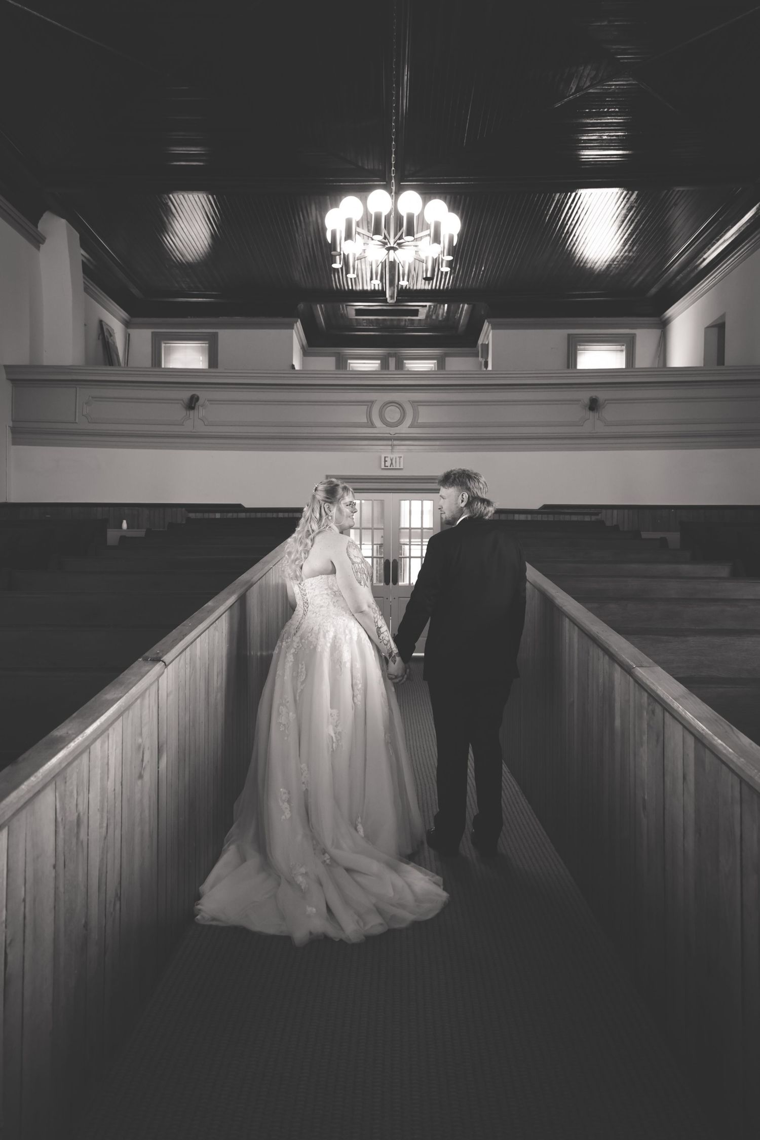 Black and white photo of bride and groom holding hands in church aisle with chandelier above.