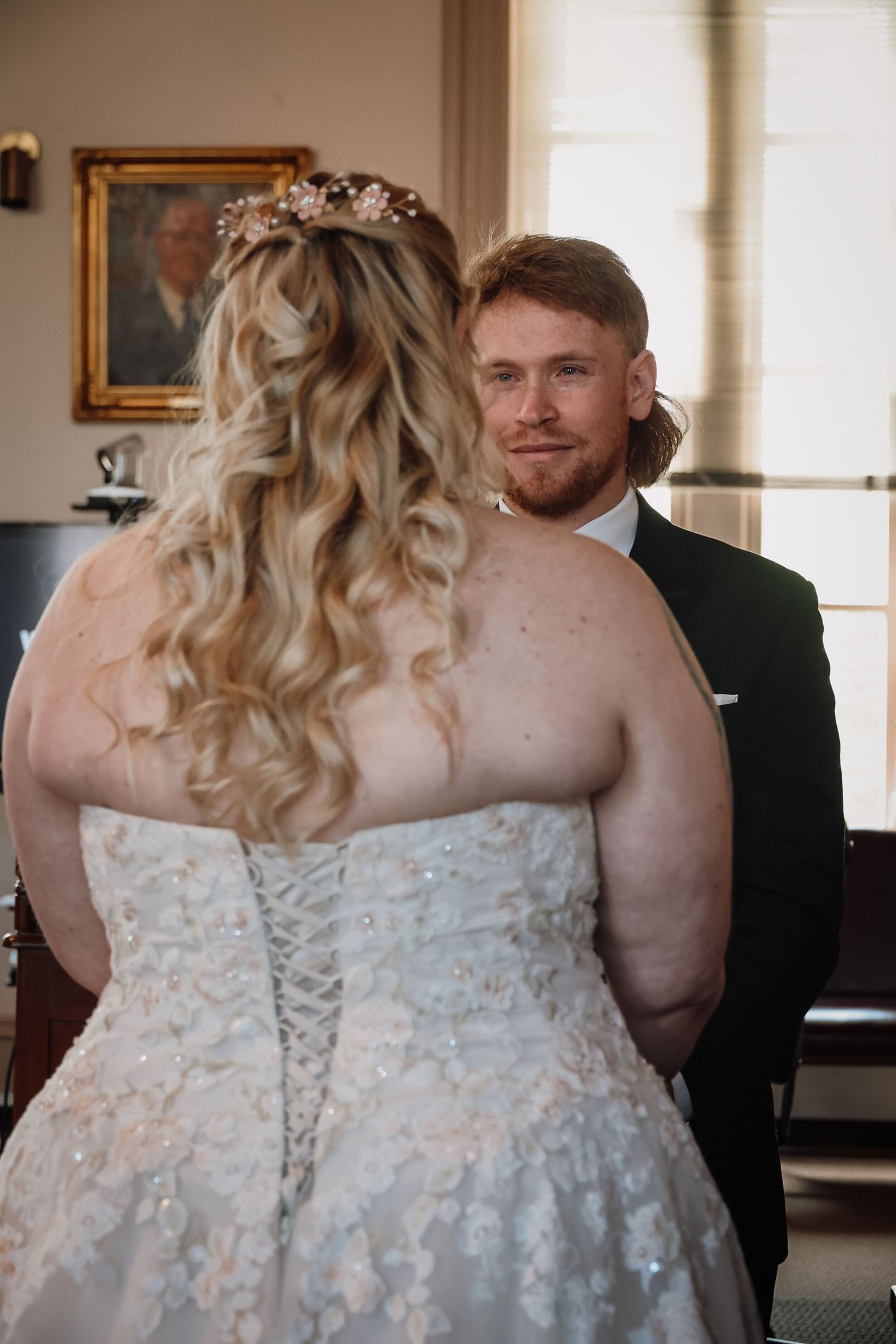 Bride and groom share an intimate moment, bride in lace corset gown with floral hair accessory, groom in dark suit.