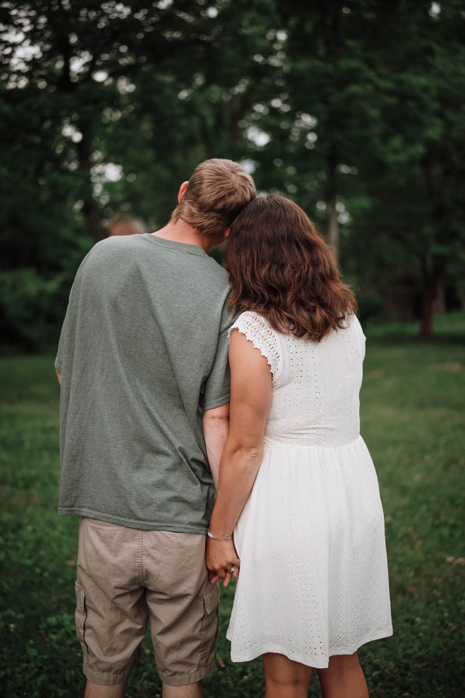 A couple holds hands from behind, standing in a lush green park, the man in a gray tee and the woman in a white dress.