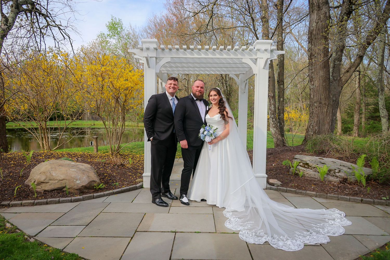 Bride and groom pose with officiant under white pergola in lush garden setting during spring wedding ceremony.