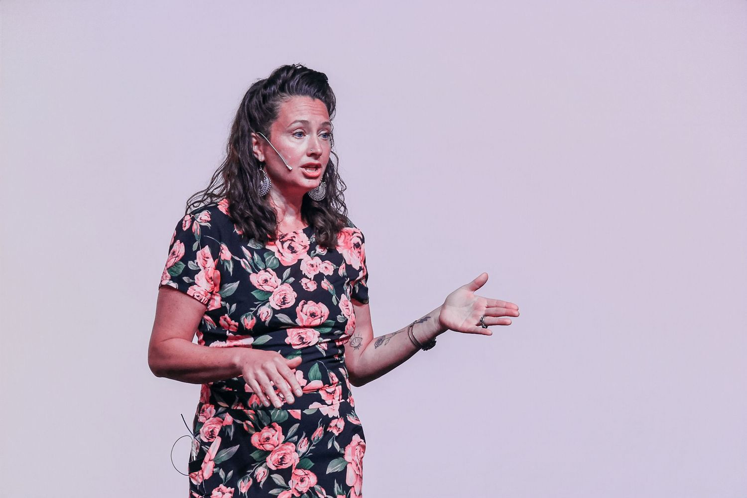 Woman in floral dress gesturing while speaking on stage against a light background.