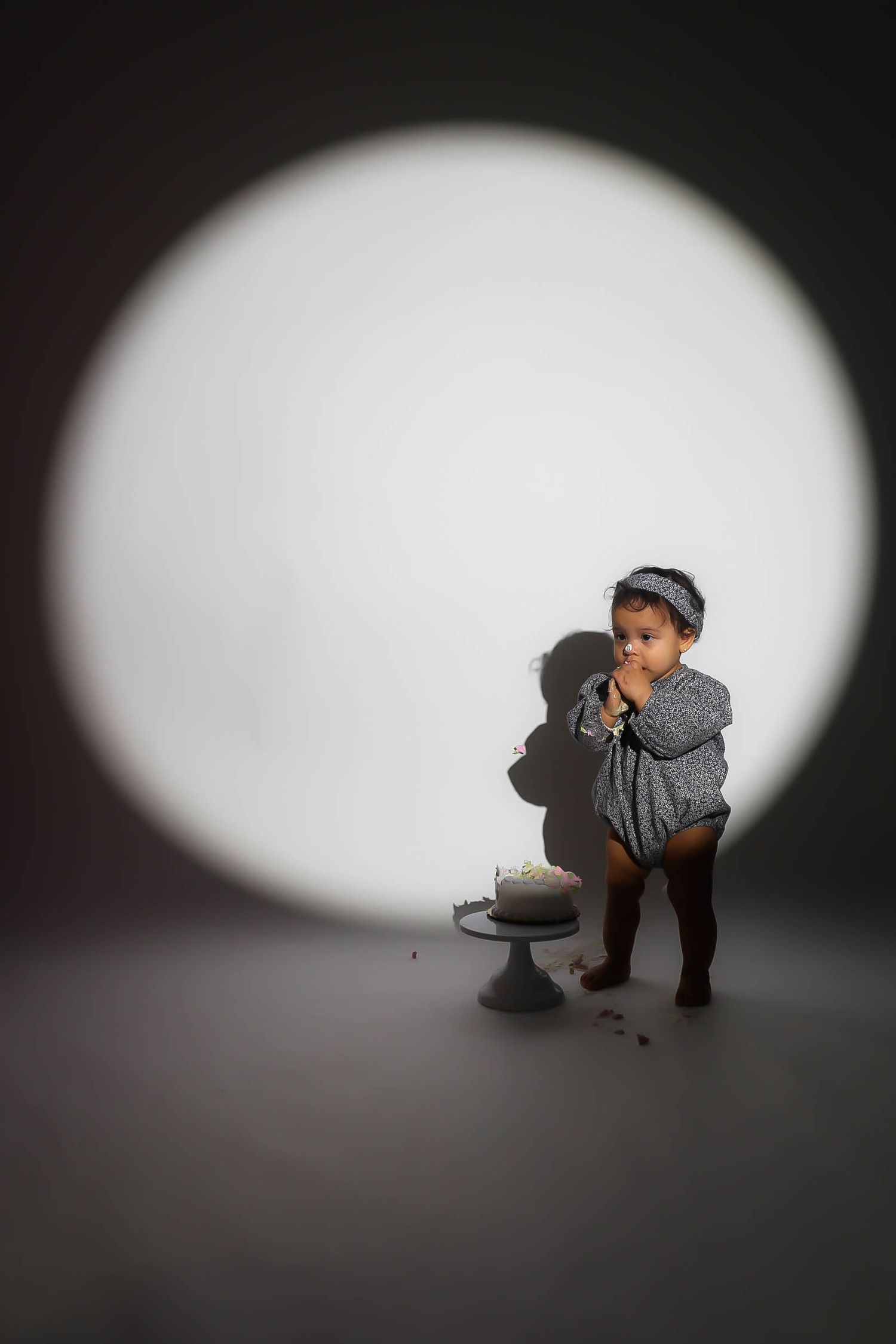 Toddler in gray romper eating cake beside a small cake stand under a dramatic circular spotlight.