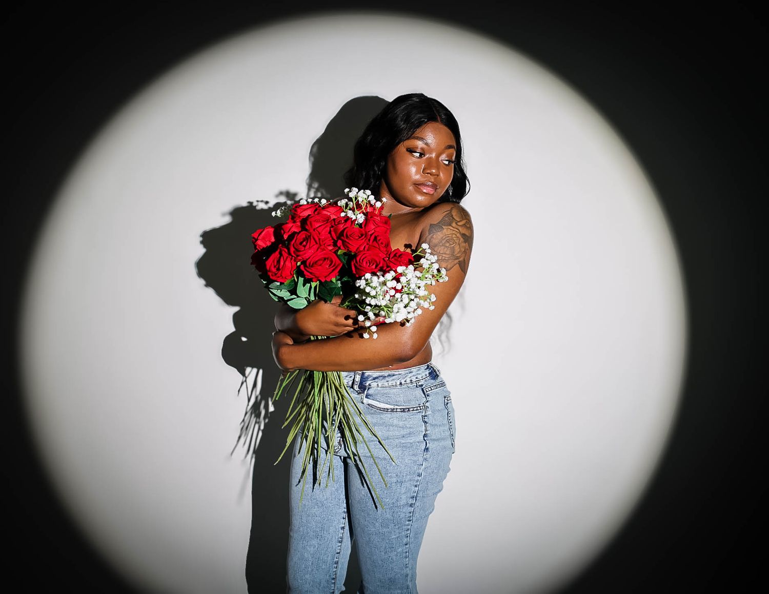 Young Black woman embracing large red roses bouquet in jeans, standing in circular spotlight on dark background.