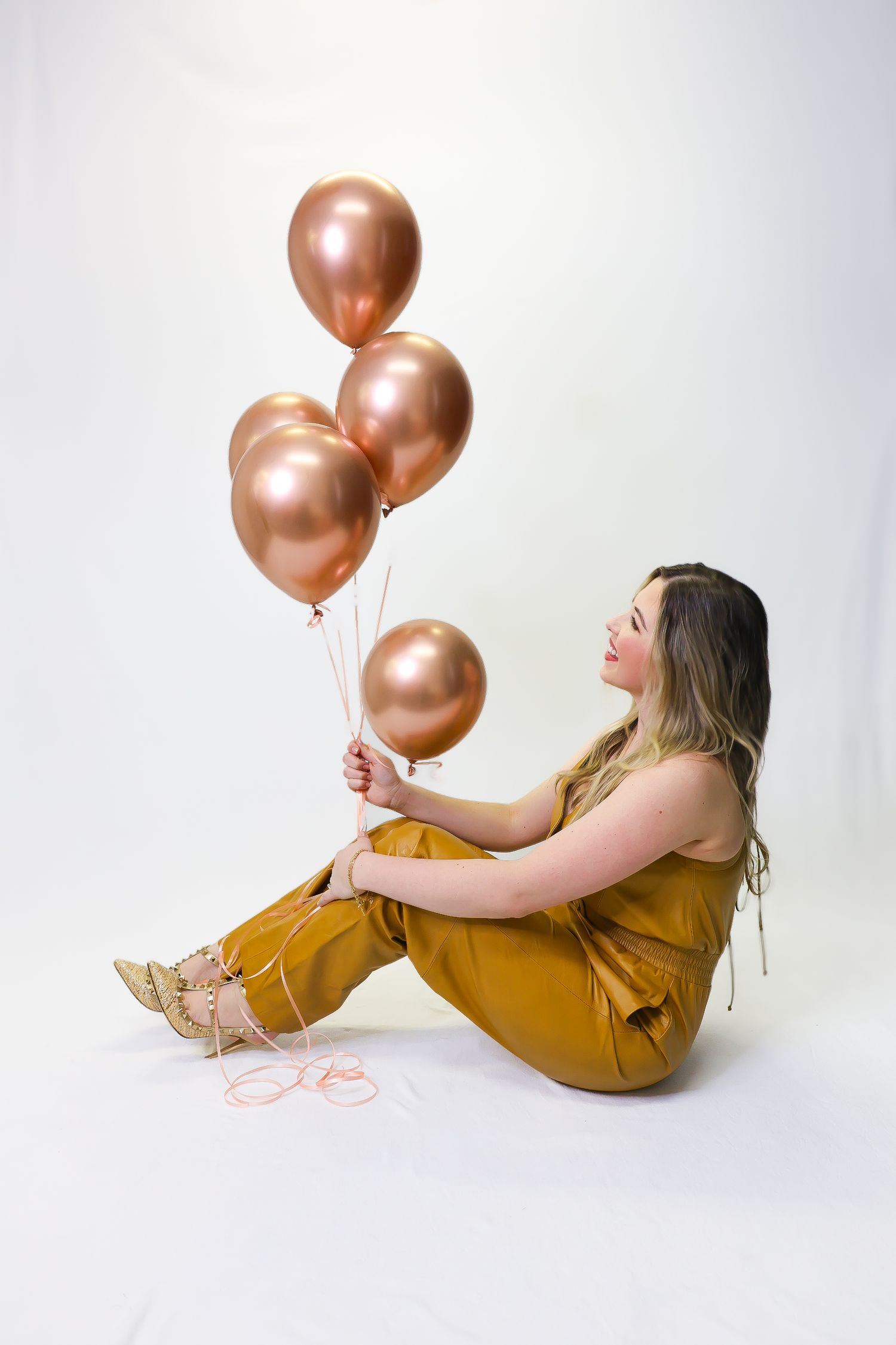 Woman in mustard jumpsuit sitting on floor holding rose gold metallic balloons against white background.