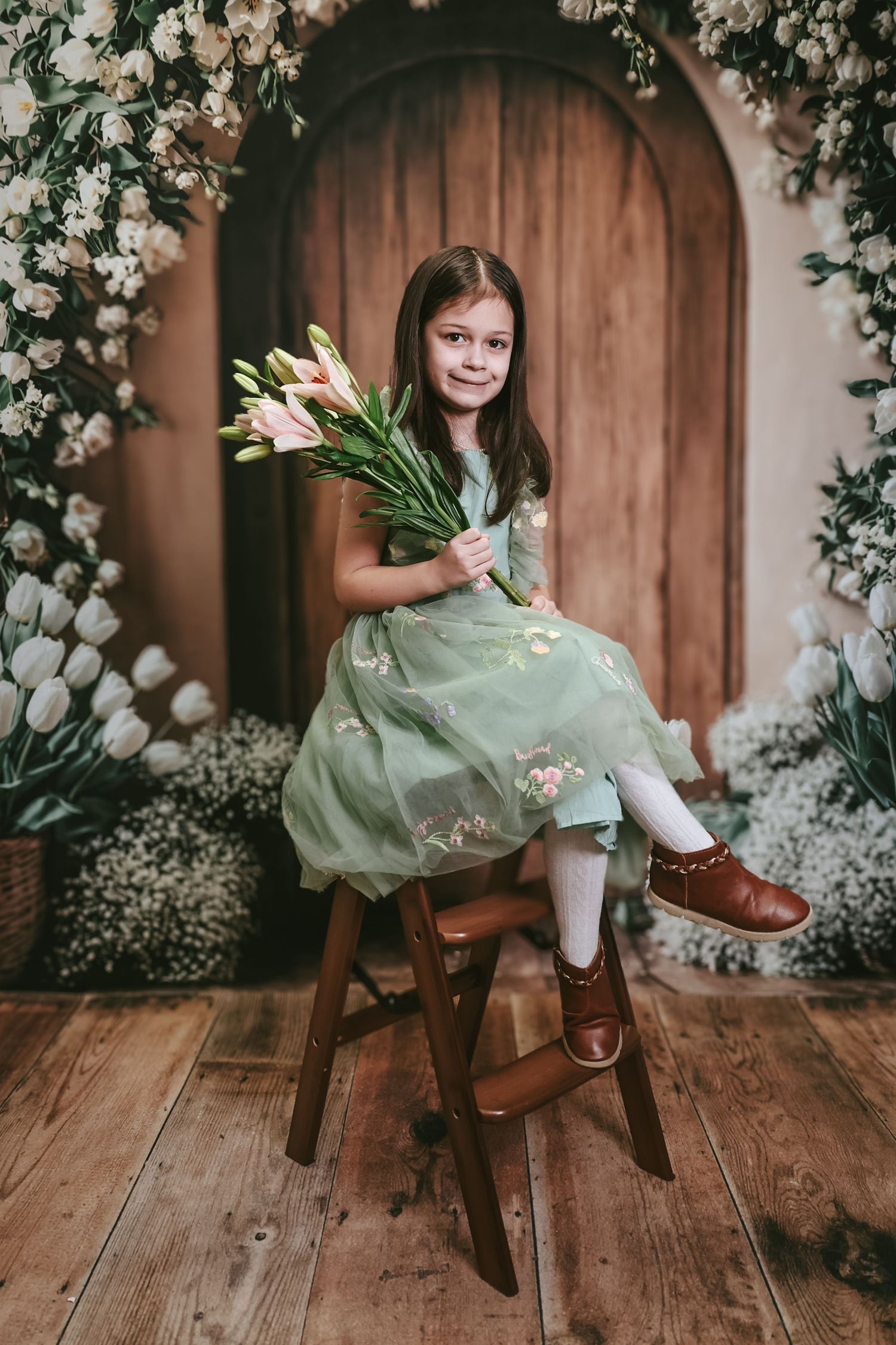 Young girl in green dress holding pink tulips sits on wooden stool surrounded by white flowers near rustic door.