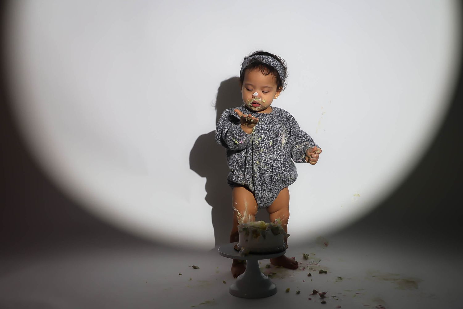 Toddler in a sparkly gray outfit sitting on a cake smash pedestal against a white studio background.