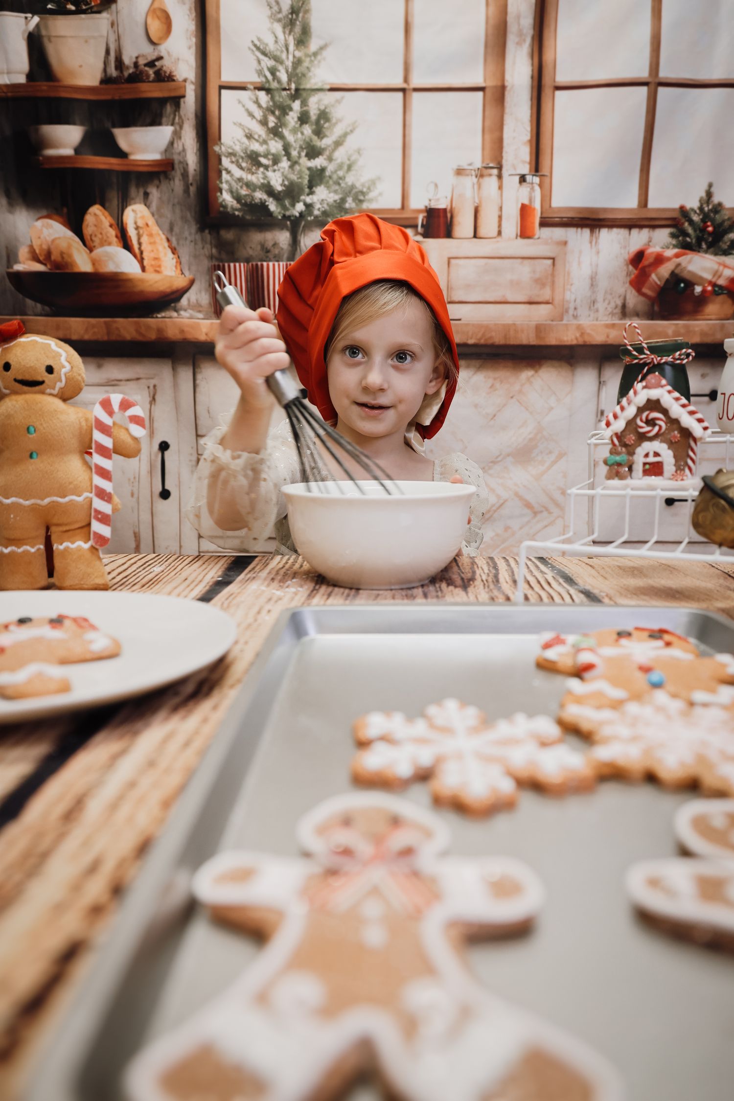 Young child in orange chef hat whisks batter surrounded by decorated Christmas gingerbread cookies on festive kitchen table.