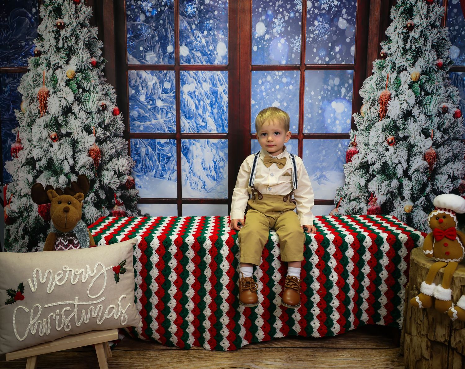 Young boy in suspenders sits on a colorful Christmas blanket between decorated trees in a festive holiday photo setting.