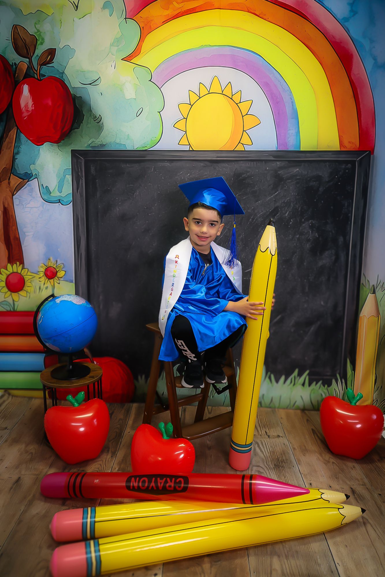 Boy in blue graduation cap and gown poses with oversized pencils and apples in a colorful school-themed photo backdrop.
