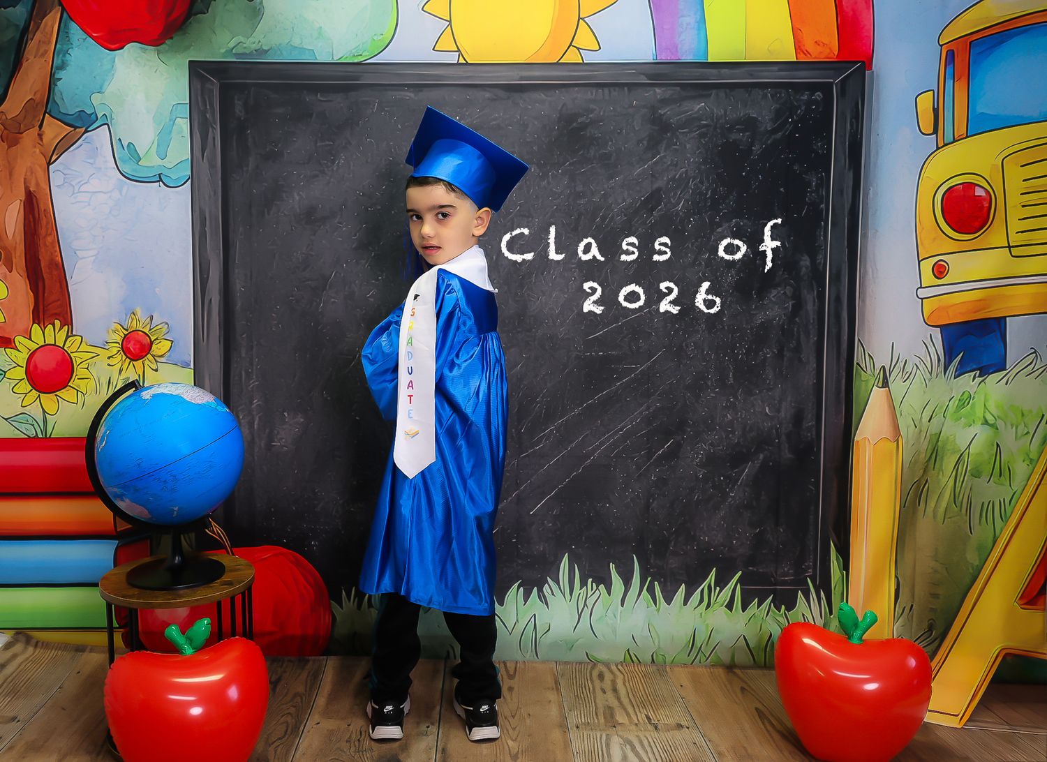 Young boy in blue graduation cap and gown poses before Class of 2026 chalkboard sign with colorful school backdrop.