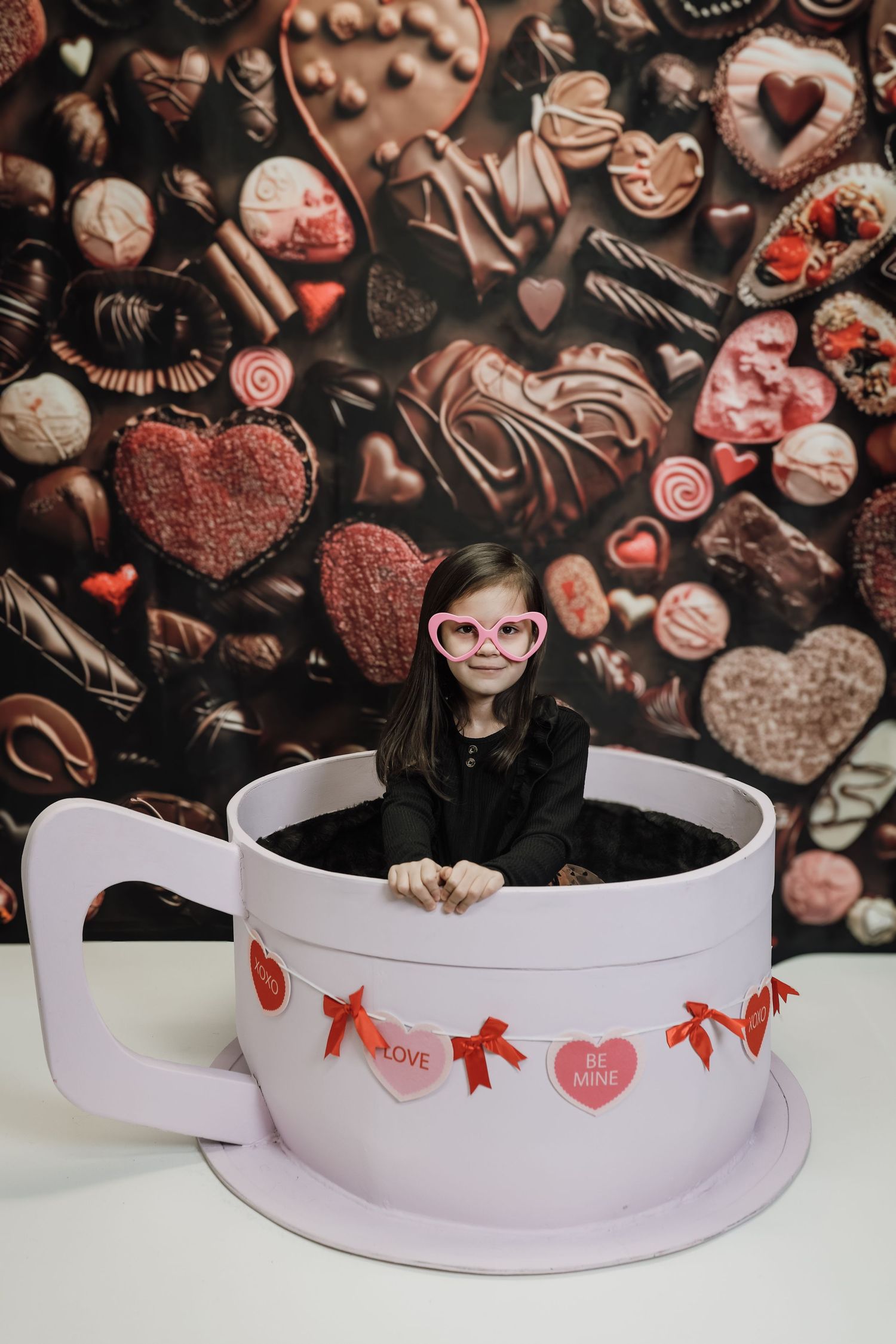 Girl in heart sunglasses sits in giant teacup with Valentine candy garland against chocolate backdrop.
