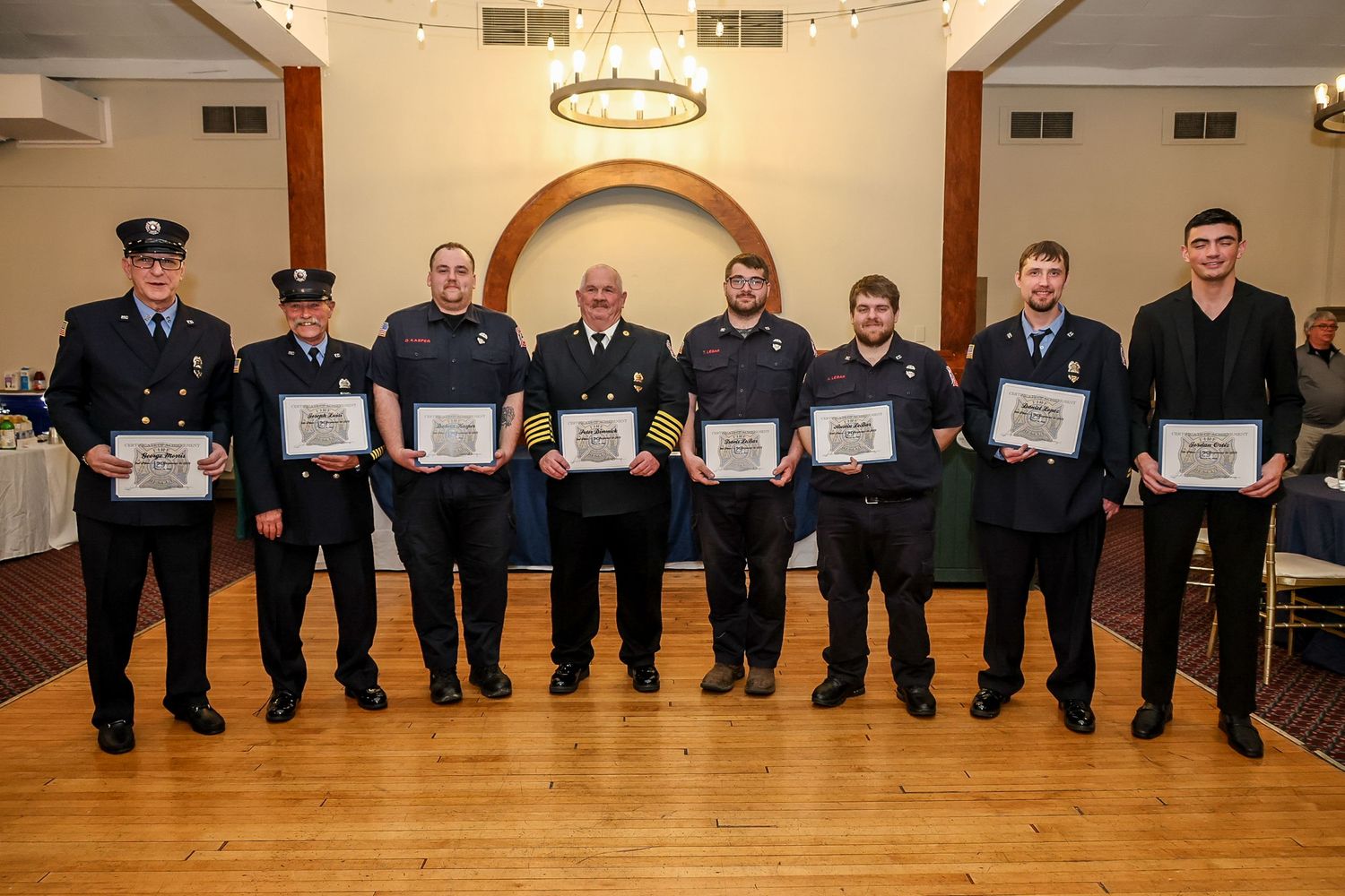 Seven firefighters in dress uniforms proudly display award certificates at a formal recognition ceremony.