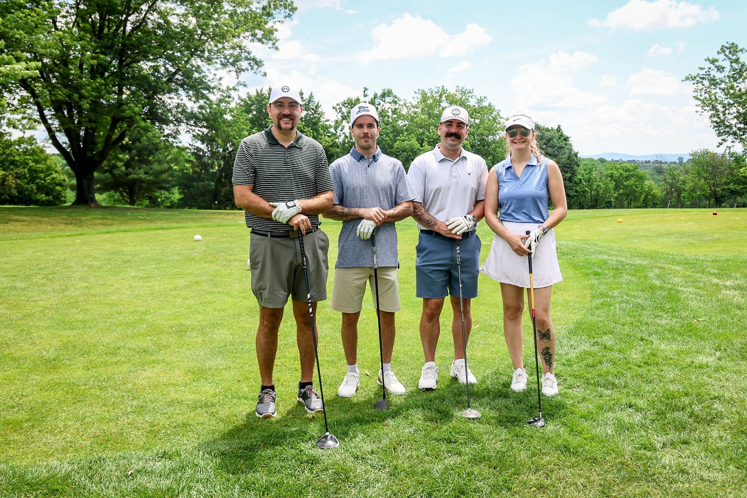 Four golfers posing together on a sunny green golf course, smiling and holding their clubs on a beautiful summer day.