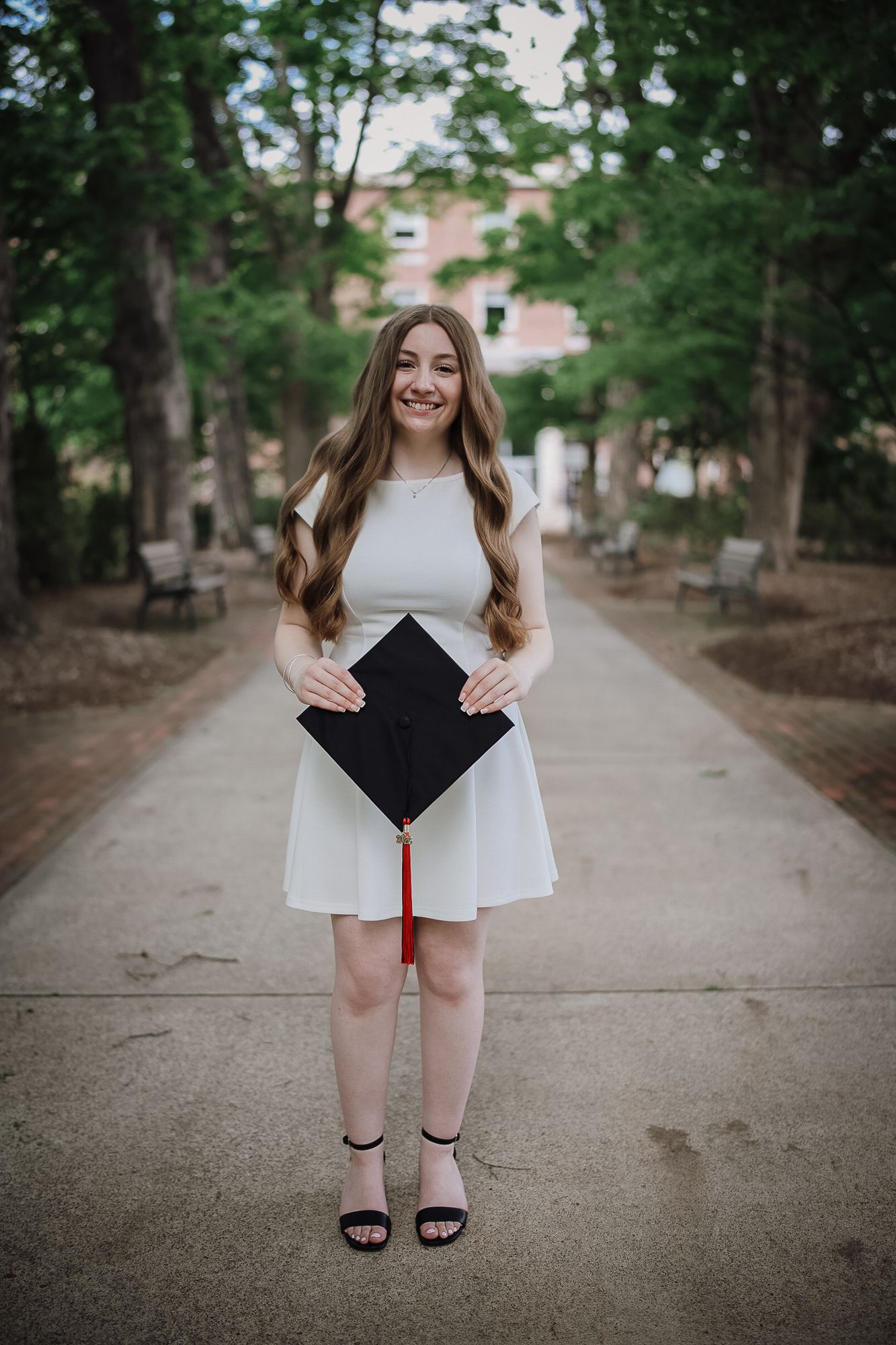 Young woman in white dress holding black graduation cap with red tassel, smiling on tree-lined campus walkway.