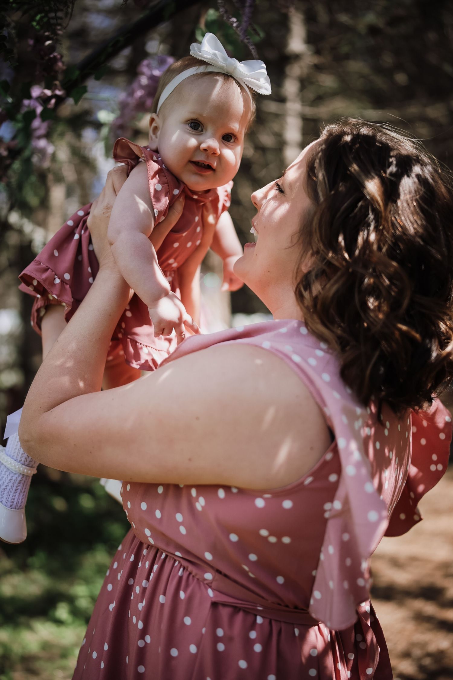 A mother in a pink polka dot dress lifts her smiling baby girl wearing a matching dress outdoors.