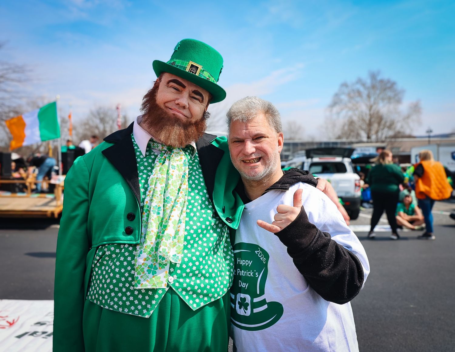 Two men pose in green St. Patrick's Day outfits, one dressed as a leprechaun, at an outdoor celebration.
