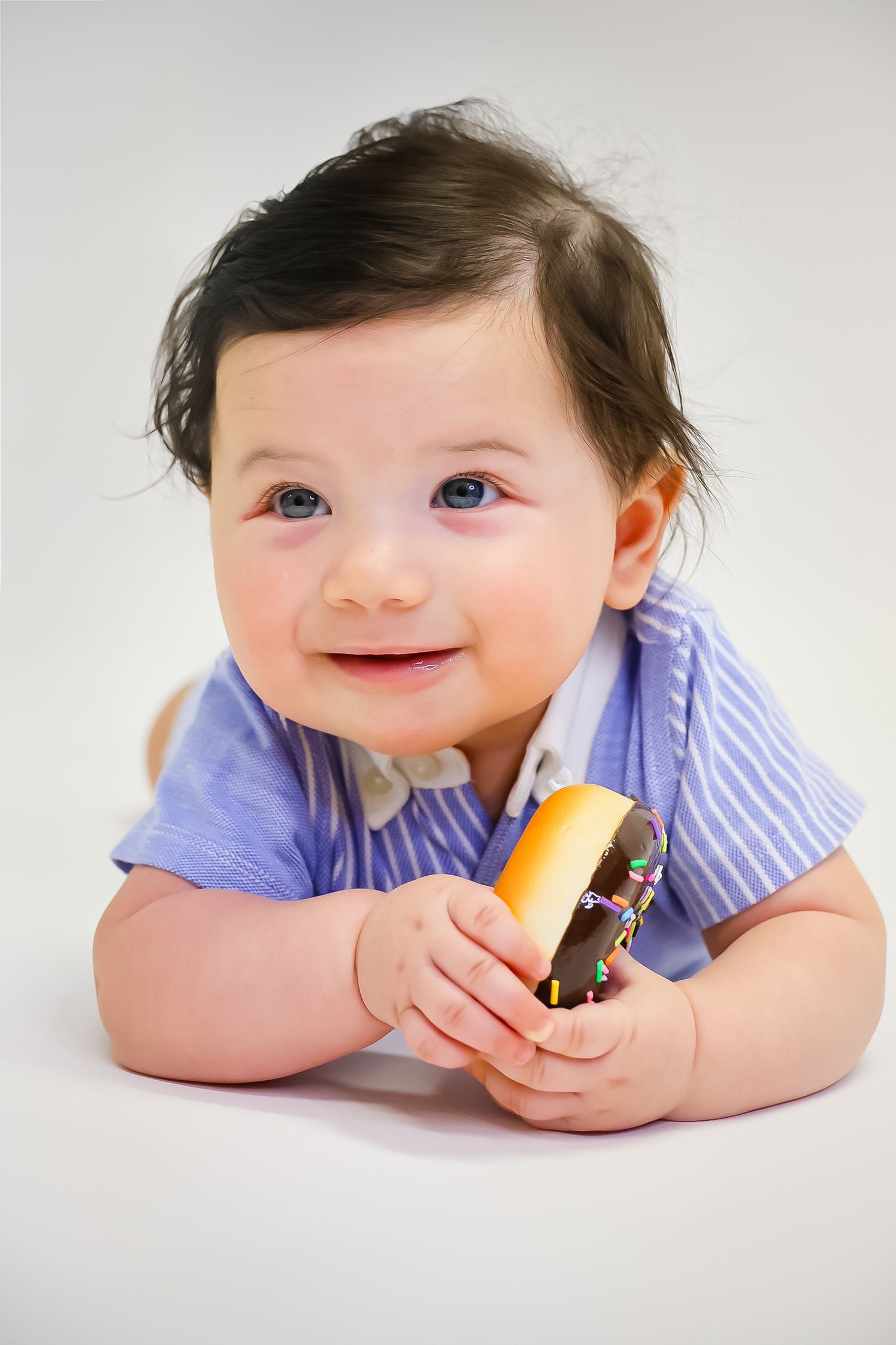 A baby in a purple shirt lies on their stomach while playfully eating a sandwich against a white background.