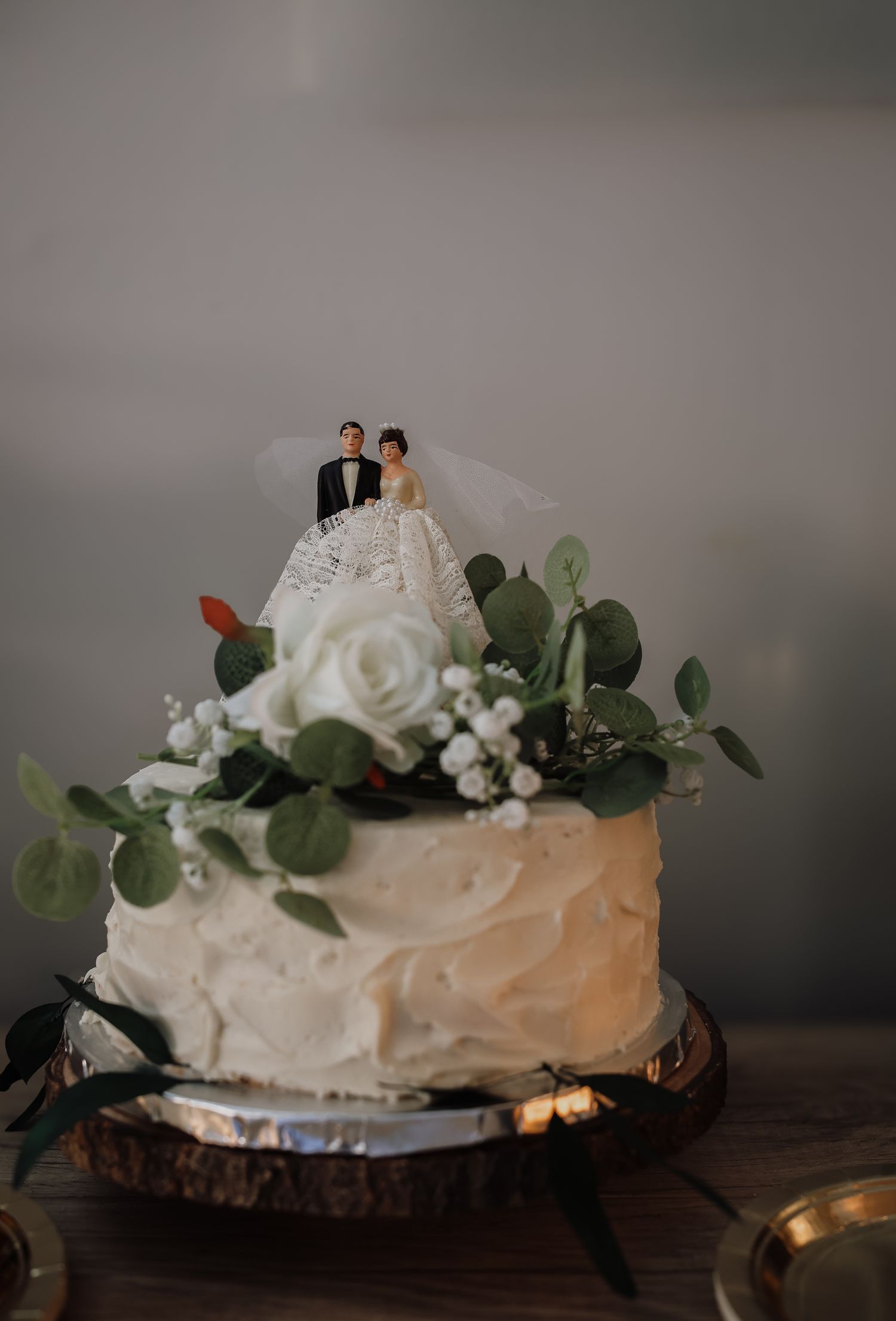 A rustic white wedding cake decorated with fresh eucalyptus leaves and roses sits on a wooden surface in moody lighting.