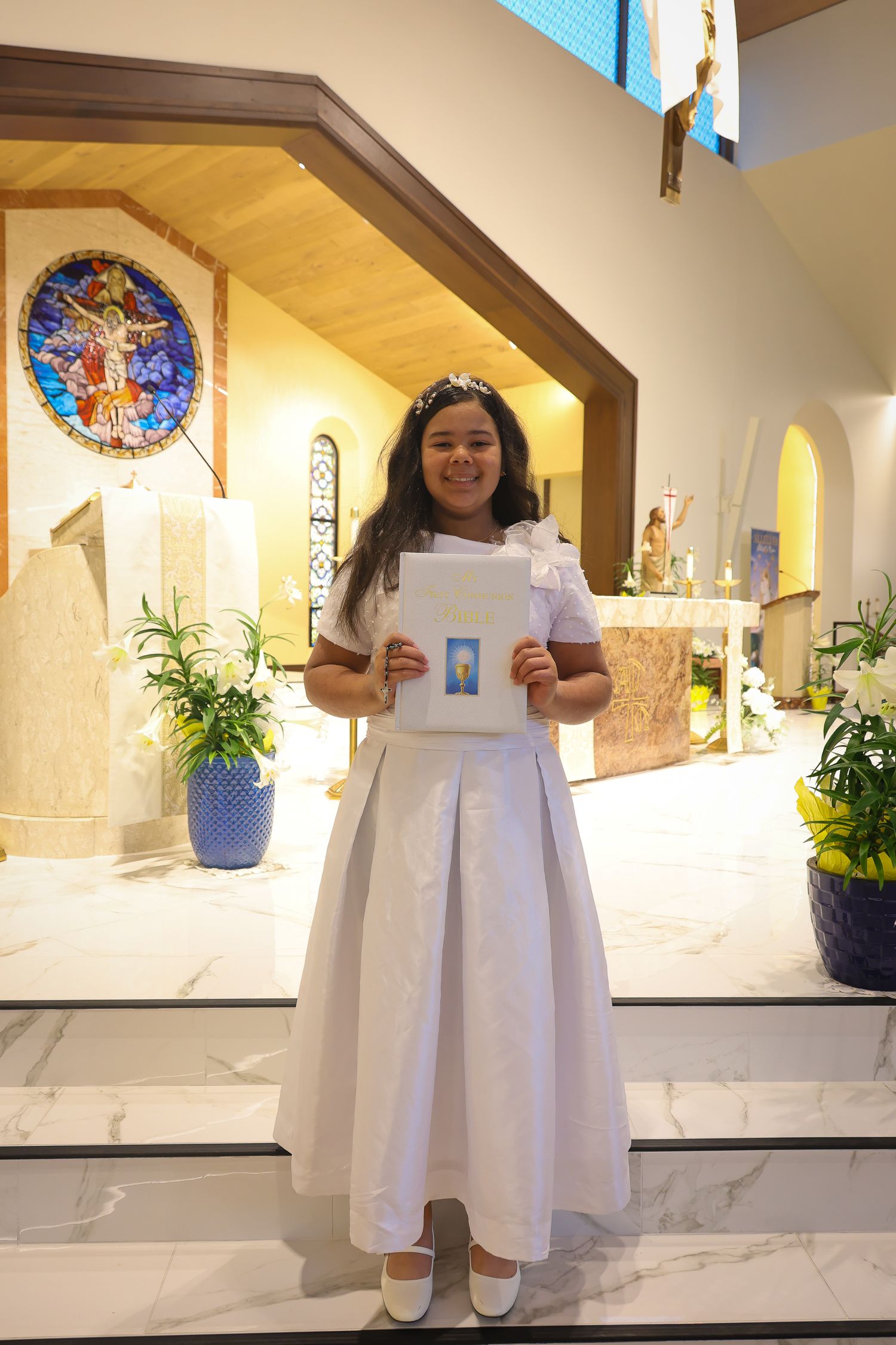 A person in a white dress stands in a church hallway with stained glass windows and potted plants.