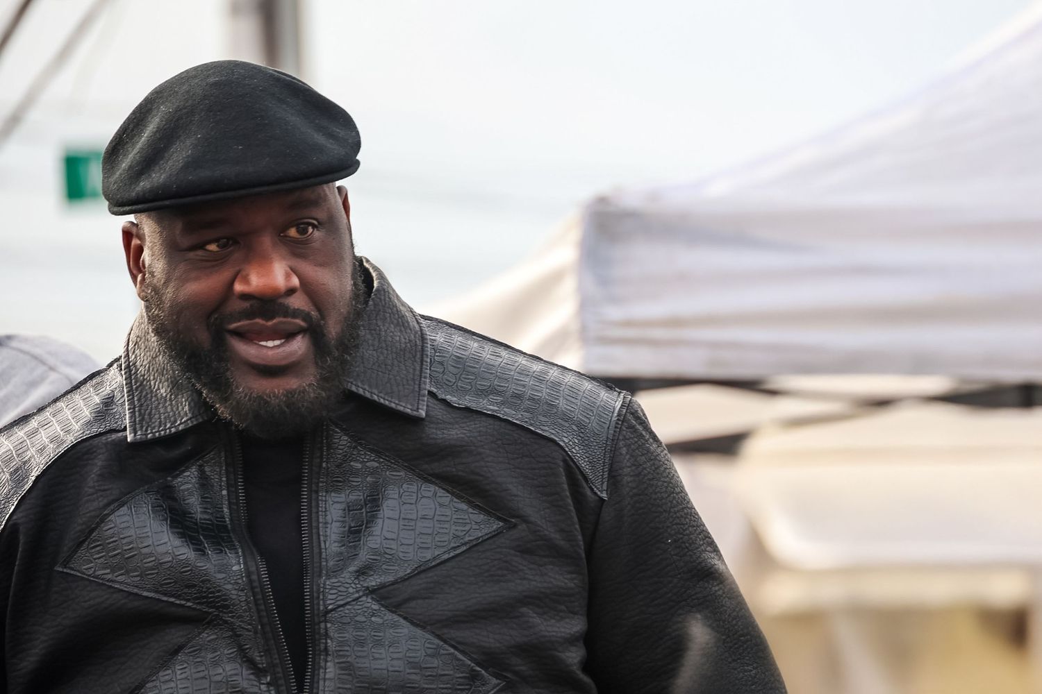 Person wearing a black leather jacket and flat cap smiles warmly while standing outdoors.